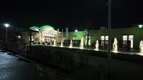 Vista-Nocturna-De-La-Entrada-Del-Baño-Termal-Arcen-Iluminada-Con-Luces-Verdes-Y-Una-Fuente-De-Agua-En-Primer-Plano.