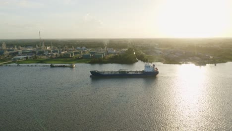 Antena:-Un-Petrolero-Navegando-Hacia-El-Muelle-En-El-Río-Surinam,-Con-Puesta-De-Sol-De-Fondo-Y-Un-Dron-Siguiéndolo