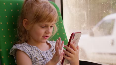 Smiling-child-girl-in-city-bus-or-tram-transport-use-mobile-cell-phone-typing-browsing-say-wow-yes