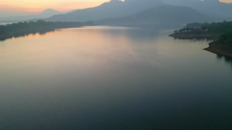 Tilt-up-shot-of-golden-sunset-over-Bhandardhara-Lake-with-the-refection-of-trees,-mountains-and-amazing-colors-on-the-water-near-Igatpuri,-in-the-western-ghats-Maharashtra-India-4K-Drone