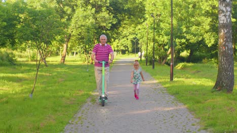 Senior-modern-man-grandfather-tourist-riding-electric-scooter-with-granddaughter-child-girl-in-park