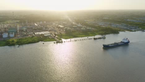 Aerial:-Oil-tanker-ship-sailing-to-dock,-Suriname-river,-sunset-in-background,-drone-ascending