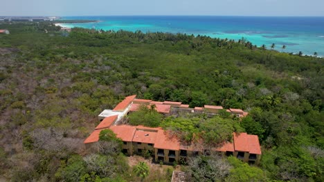 Abandoned-beach-hotel-damaged-by-hurricane-on-tropical-coastline--overgrown-with-trees,-aerial-view