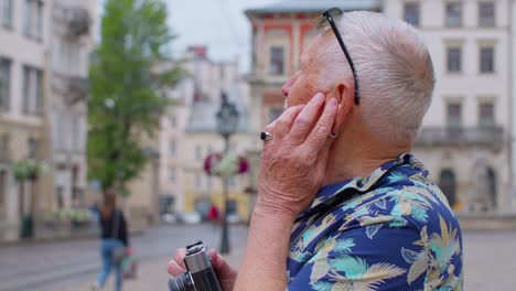 Senior-tourist-with-retro-photo-camera-listening-to-music-on-earphones-dancing-outdoors-having-fun