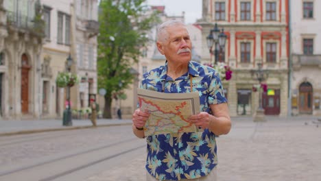 Elderly-stylish-tourist-grandfather-man-walking-along-street-looking-for-way-using-paper-map-in-city