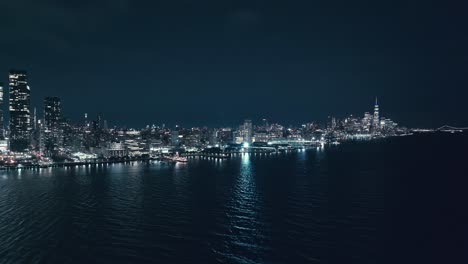 Eine-Drohne-Umkreist-Den-East-River,-Während-Die-Lichter-Des-Hafens-Und-Der-Promenade-Unter-Der-Skyline-Von-New-York-Auf-Dem-Wasser-Funkeln