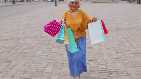 Senior-stylish-tourist-woman-walking-along-street-in-city-center-with-bags-after-shopping-in-mall