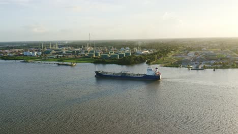 Vista-Aérea:-Un-Petrolero-Navegando-Hacia-El-Muelle-En-El-Río-Surinam,-Con-Una-Refinería-Al-Fondo-Y-Un-Dron-Siguiéndolo