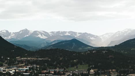 Drone-rises-above-Estes-Park-Colorado-as-sunlight-breaks-between-clouds-across-snow-covered-Rocky-Mountains