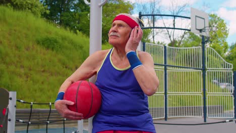 Abuelo-Anciano-Después-Del-Entrenamiento-Deportivo-De-Baloncesto-Sentado,-Escuchando-Música-En-El-Patio-De-Recreo