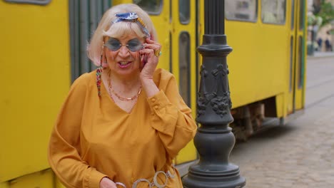 Senior-old-tourist-woman-in-stylish-clothes-talking-on-mobile-phone-while-walking-on-city-street