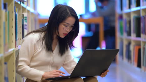 Portrait-of-happy-joyful-pretty-female-student-busy-hard-working-sitting-against-background-of-bookshelves-in-university-library-holding-laptop-and-backpack-looking-to-the-side,-smiling-pleasant