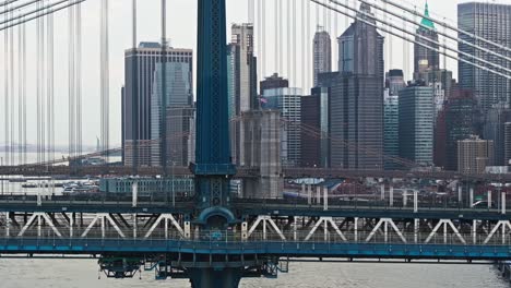 Drone-pans-across-Manhattan-bridge-with-cars-crossing,-NYC-skyline-buildings-and-Brooklyn-bridge-behind-cable-wires