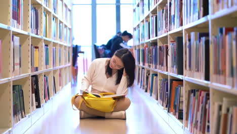 Portrait-of-happy-joyful-pretty-female-student-busy-hard-working-sitting-against-background-of-bookshelves-in-university-library-holding-laptop-and-backpack-looking-to-the-side,-smiling-pleasant