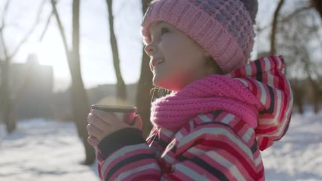 Child-girl-kid-drinking-hot-drink-tea-from-cup-on-snowy-road-in-winter-park,-Christmas-holidays