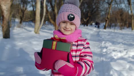 Cheerful-smiling-child-girl-looking-at-camera-with-Christmas-present-gift-box-in-winter-snowy-park