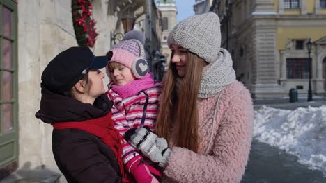 Travelers-sisters-holding-younger-sister-child-girl-in-hands,-talking,-smiling,-embracing-in-city