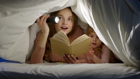 Mother-and-daughter-lying-in-bed-at-home-under-duvet-blanket-and-reading-bedtime-stories,-fairytales