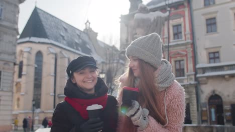 Two-smiling-women-tourists-traveling-together,-drinking-hot-tea,-coffee-from-cups-on-city-street