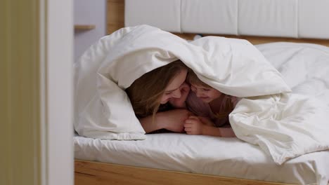 Mother-lying-on-bed-under-duvet-blanket-with-daughter-kid-telling-secrets-to-each-other,-smilling