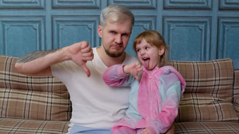 Displeased-father-and-child-daughter-kid-in-pajamas-sit-on-couch-in-room,-showing-thumb-down-dislike