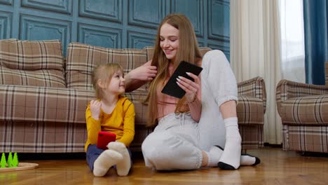 Mother-and-daughter-child-sit-on-floor-at-home-working-on-mobile-phone-and-tablet,-watching-cartoon