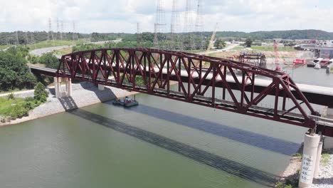 Railroad-through-truss-bridge-above-river-water-with-barges-docked-at-port-in-background,-Kentucky,-circle-aerial