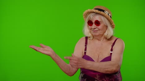 Senior-old-woman-tourist-in-swimsuit-points-fingers-on-left-at-blank-space-shows-advertising-content