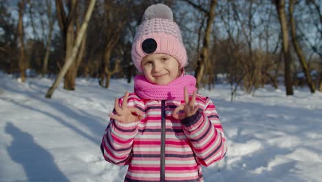 Child-girl-smiling,-showing-ok-sign,-thumbs-up-gesture-on-snowy-road-in-winter-sunny-park-forest