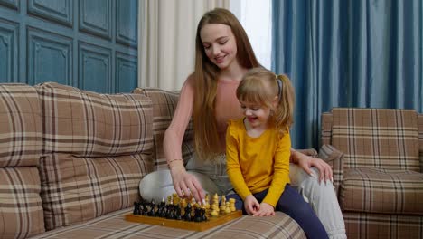 Feliz-Joven-Madre-Enseñando-A-Su-Hija-Pequeña-A-Jugar-Ajedrez-En-Una-Tabla-De-Madera-En-El-Sofá-De-Casa