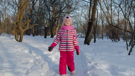 Child-girl-walking-on-snowy-road,-fooling-around,-smiling,-looking-at-camera-in-winter-park-forest