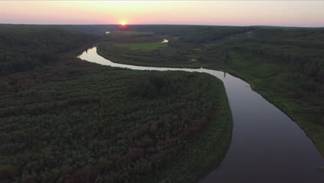 A-drone-flies-backwards-over-a-calm,-winding-river-valley-at-sunset