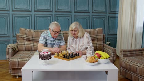 Retired-senior-couple-talking-drinking-tea,-playing-chess-in-modern-living-home-room-lounge-together