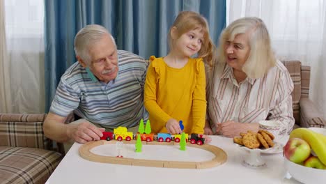 Child-girl-kid-involved-in-build-board-game-ride-toy-train-on-railroad-with-senior-grandparents-home