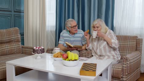 Retired-senior-couple-talking-drinking-tea,-reading-book-in-modern-living-home-room-lounge-together