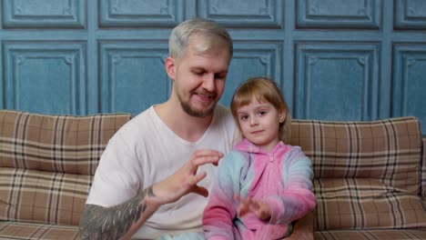 Father-and-child-daughter-kid-in-pajamas-sit-on-couch-smiling,-making-sign-of-shape-heart-near-chest