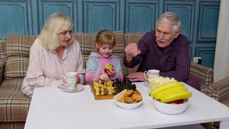 Senior-grandparents-and-child-granddaughter-spending-time-home-together,-sitting,-playing-chess-game