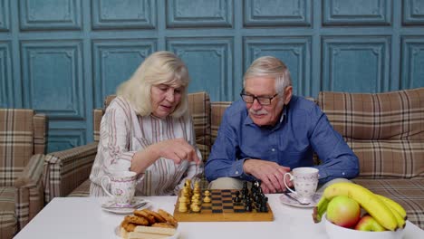 Una-Pareja-De-Ancianos-Jubilados-Hablando-Bebiendo-Té,-Jugando-Al-Ajedrez-En-Un-Moderno-Salón-De-Estar-Juntos