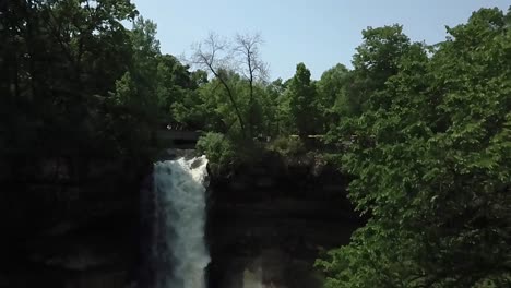 Wide-aerial-shot-moving-upwards-while-looking-at-the-Minnehaha-waterfall-with-its-rushing,-white-water-surrounded-by-large,-green-trees