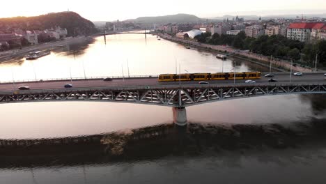 Drohne-Folgt-Der-Gelben-Straßenbahn-In-Budapest,-Petőfi-Brücke-Im-Wunderschönen-Sonnenuntergang-über-Der-Donau