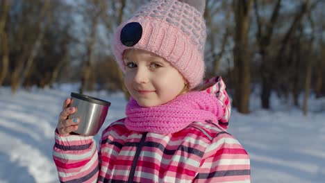 Child-girl-kid-drinking-hot-drink-tea-from-cup-on-snowy-road-in-winter-park,-Christmas-holidays