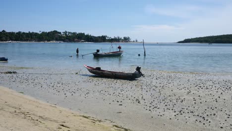 Long-tail-boat-floating-in-shallow,-calm-sea-off-Samui-Island,-Thailand,-with-a-tourist-in-background-paddling