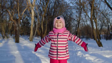 Joyful-little-child-girl-throwing-snow-up,-smiling,-playing,-showing-thumbs-up-in-winter-park-forest