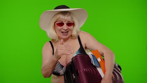 Senior-woman-tourist-in-swimsuit-with-luggage,-passport,-tickets-asking-to-follow-join-on-chroma-key