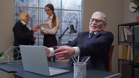 Cheerful-mature-old-businessman-raising-hands-celebrating-sudden-victory-with-colleagues-in-office