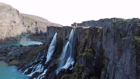 Vista-Aérea-De-Una-Figura-Masculina-De-Pie-Sobre-Un-Profundo-Y-Empinado-Cañón-En-La-Cima-De-Majestuosas-Cascadas-En-Las-Tierras-Altas-De-Islandia