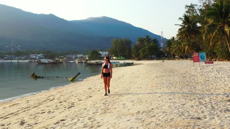 Attractive-young-woman-in-black-swimsuit-with-headphones-listening-music-walking-on-white-sandy-beach-alongside-calm-sea-in-Thailand