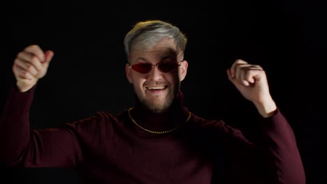 Handsome-man-in-stylish-sunglasses-dancing-to-music-rhythmically,-moving-hands-on-black-background