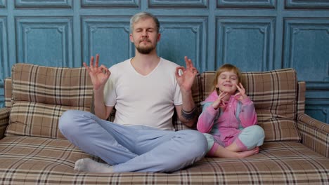 Father-and-child-kid-daughter-are-meditating-while-sitting-in-yoga-lotus-pose-on-sofa-at-home-room