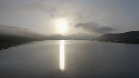 Volar-Sobre-Una-Bahía-Con-Agua-Tranquila,-El-Sol-Reflejándose-En-La-Superficie-Del-Agua-Y-Una-Densa-Nube-De-Niebla-Proveniente-De-Las-Colinas,-Al-Atardecer